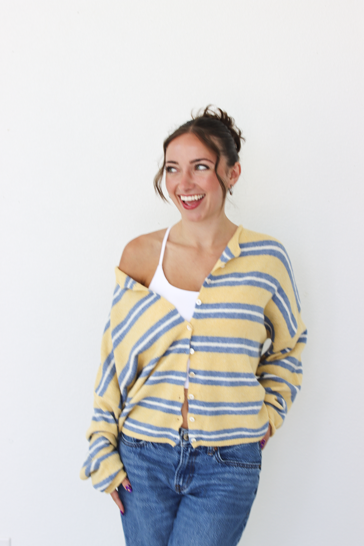 A woman wearing a yellow and blue striped knit cardigan over a white tank top and blue jeans, standing against a plain white wall.