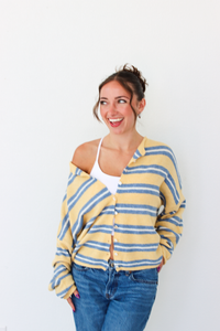 A woman wearing a yellow and blue striped knit cardigan over a white tank top and blue jeans, standing against a plain white wall.