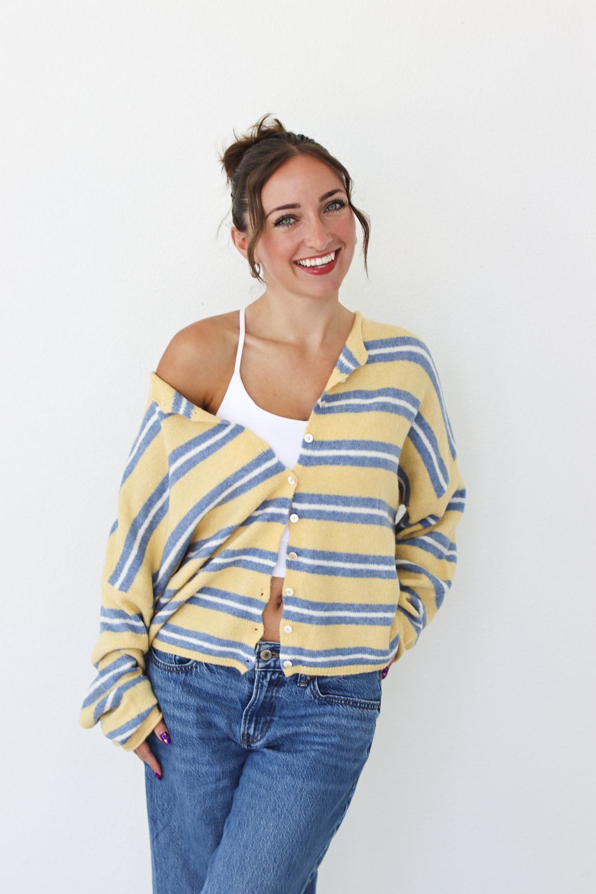 A woman wearing a yellow and blue striped knit cardigan over a white tank top and blue jeans, standing against a plain white wall.