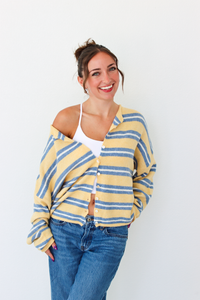 A woman wearing a yellow and blue striped knit cardigan over a white tank top and blue jeans, standing against a plain white wall.