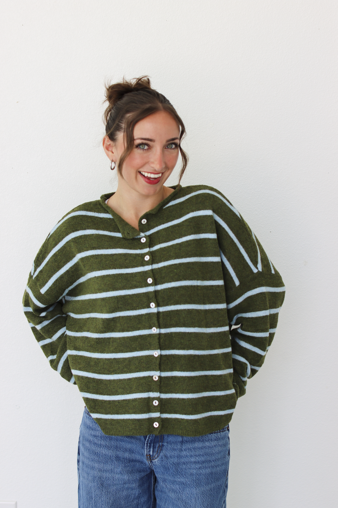 A woman wearing an olive green and light blue striped button-up cardigan sweater with blue jeans, standing against a plain white wall.