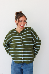 A woman wearing an olive green and light blue striped button-up cardigan sweater with blue jeans, standing against a plain white wall.