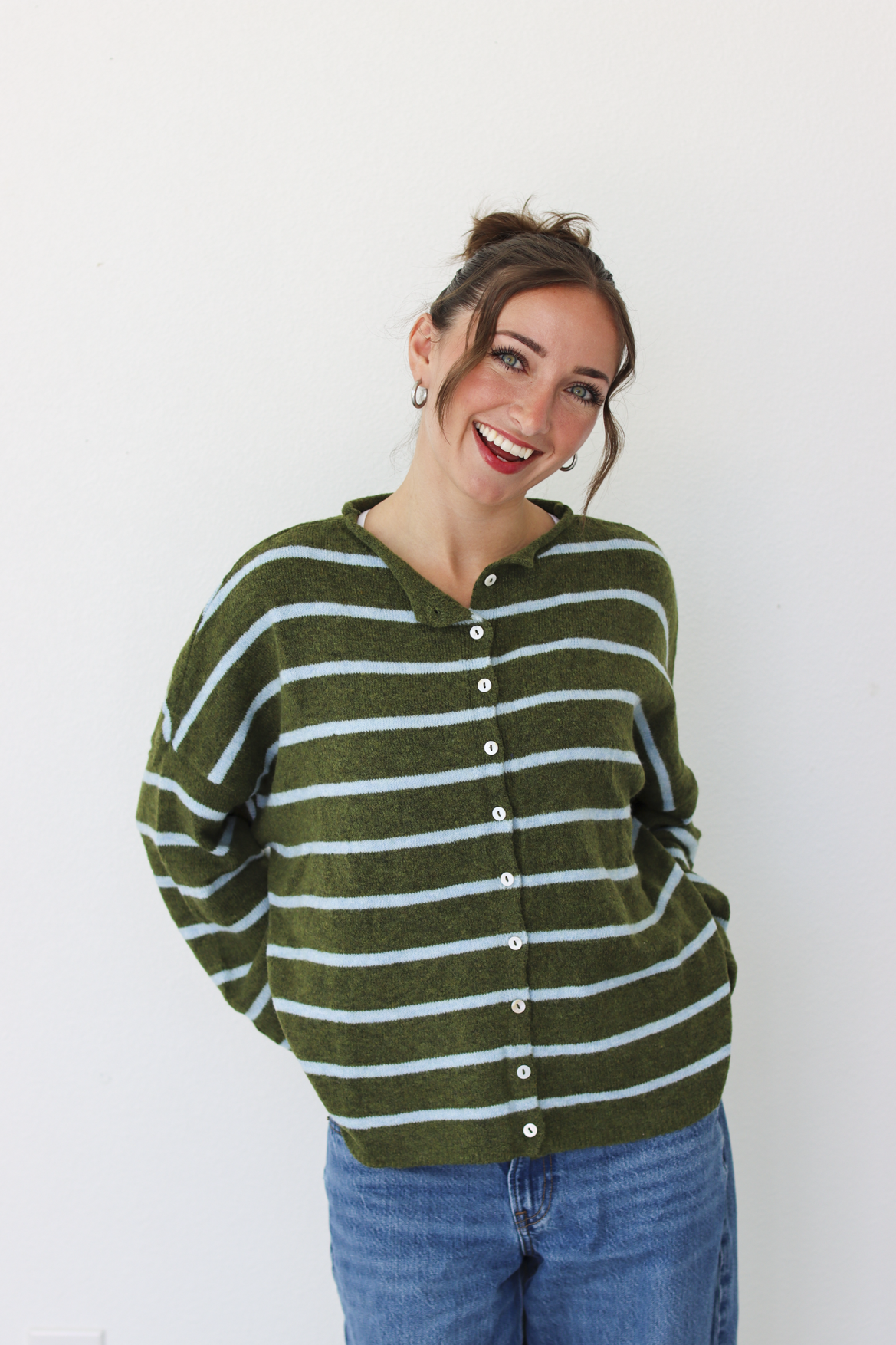 A woman wearing an olive green and light blue striped button-up cardigan sweater with blue jeans, standing against a plain white wall.