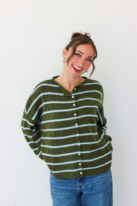 A woman wearing an olive green and light blue striped button-up cardigan sweater with blue jeans, standing against a plain white wall.