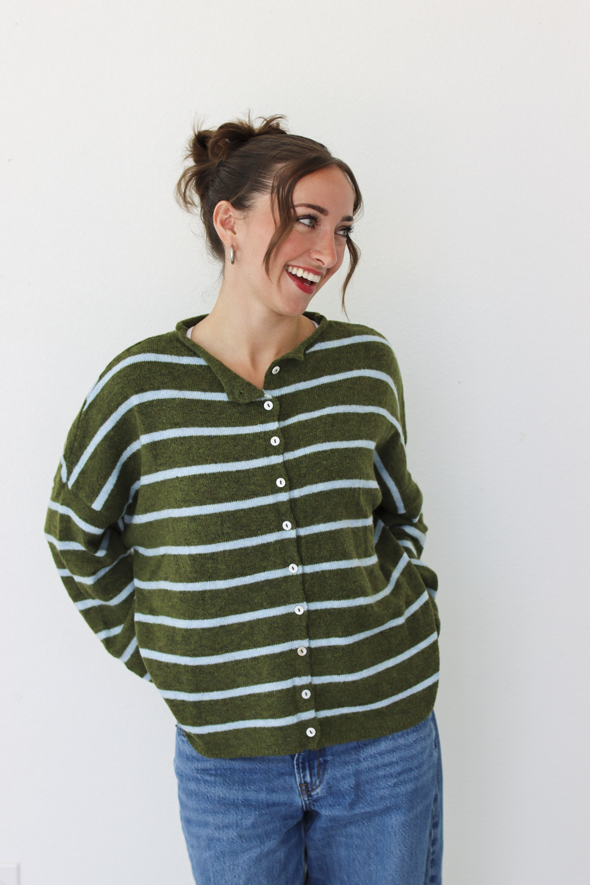 A woman wearing an olive green and light blue striped button-up cardigan sweater with blue jeans, standing against a plain white wall.