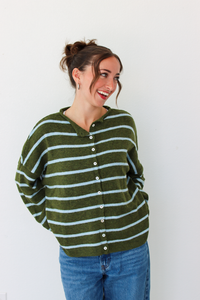 A woman wearing an olive green and light blue striped button-up cardigan sweater with blue jeans, standing against a plain white wall.