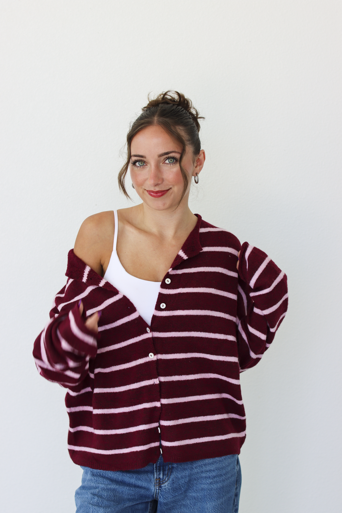 A woman wearing a burgundy and pink horizontally striped button-up cardigan over a white tank top and blue jeans, standing against a plain white wall.
