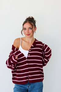 A woman wearing a burgundy and pink horizontally striped button-up cardigan over a white tank top and blue jeans, standing against a plain white wall.