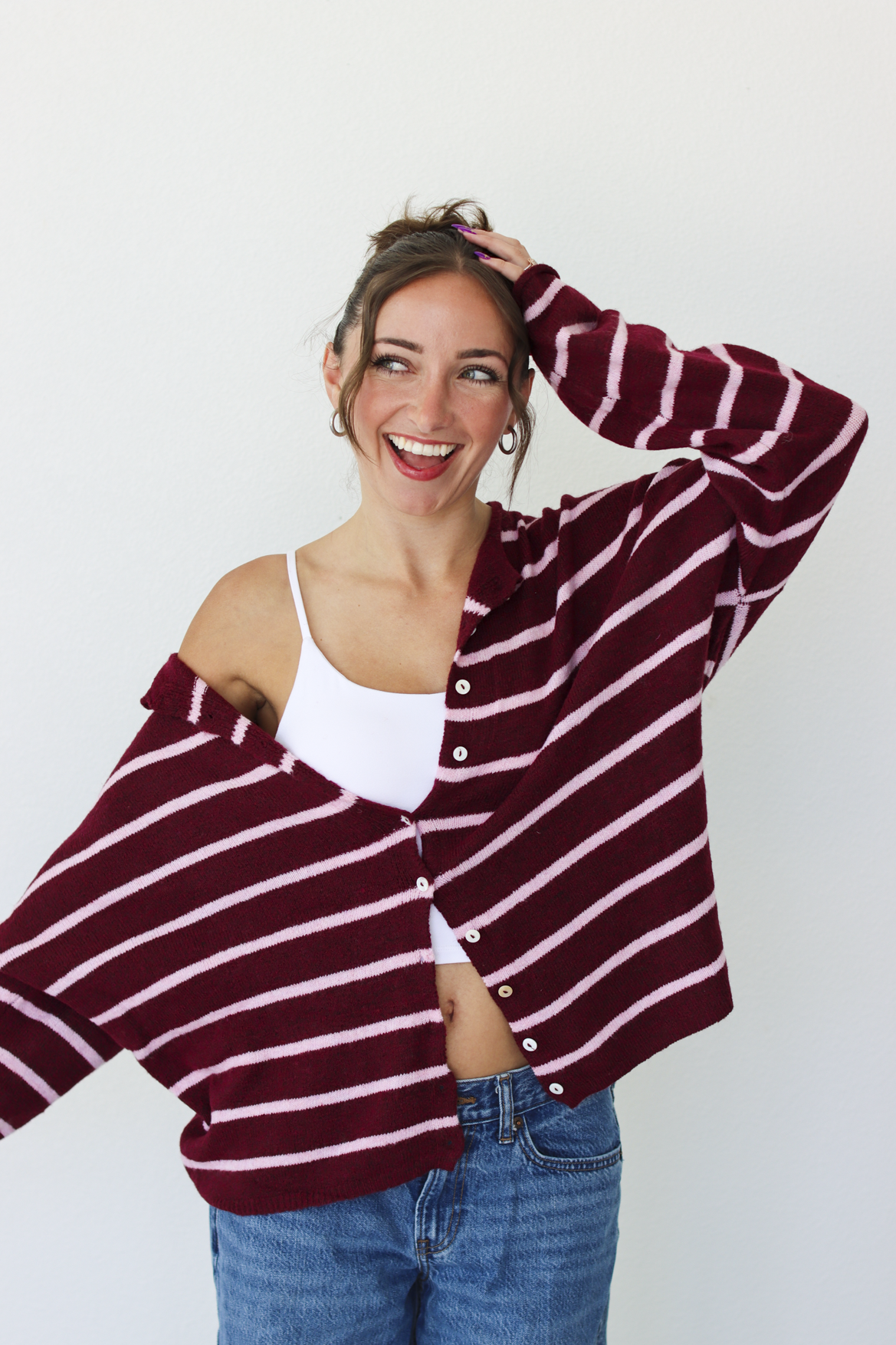 A woman wearing a burgundy and pink horizontally striped button-up cardigan over a white tank top and blue jeans, standing against a plain white wall.
