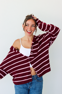 A woman wearing a burgundy and pink horizontally striped button-up cardigan over a white tank top and blue jeans, standing against a plain white wall.