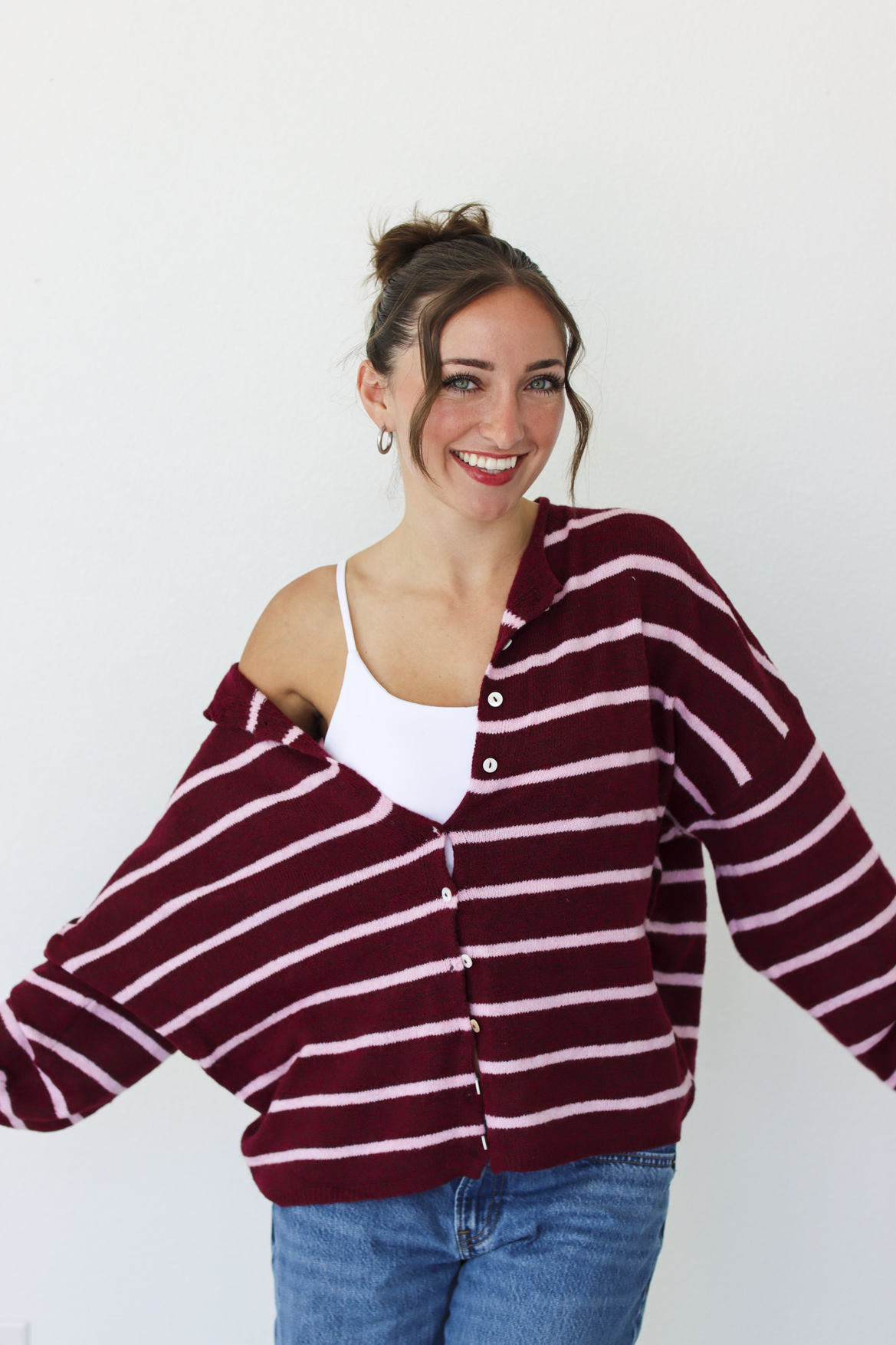 A woman wearing a burgundy and pink horizontally striped button-up cardigan over a white tank top and blue jeans, standing against a plain white wall.