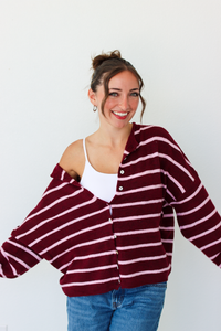 A woman wearing a burgundy and pink horizontally striped button-up cardigan over a white tank top and blue jeans, standing against a plain white wall.