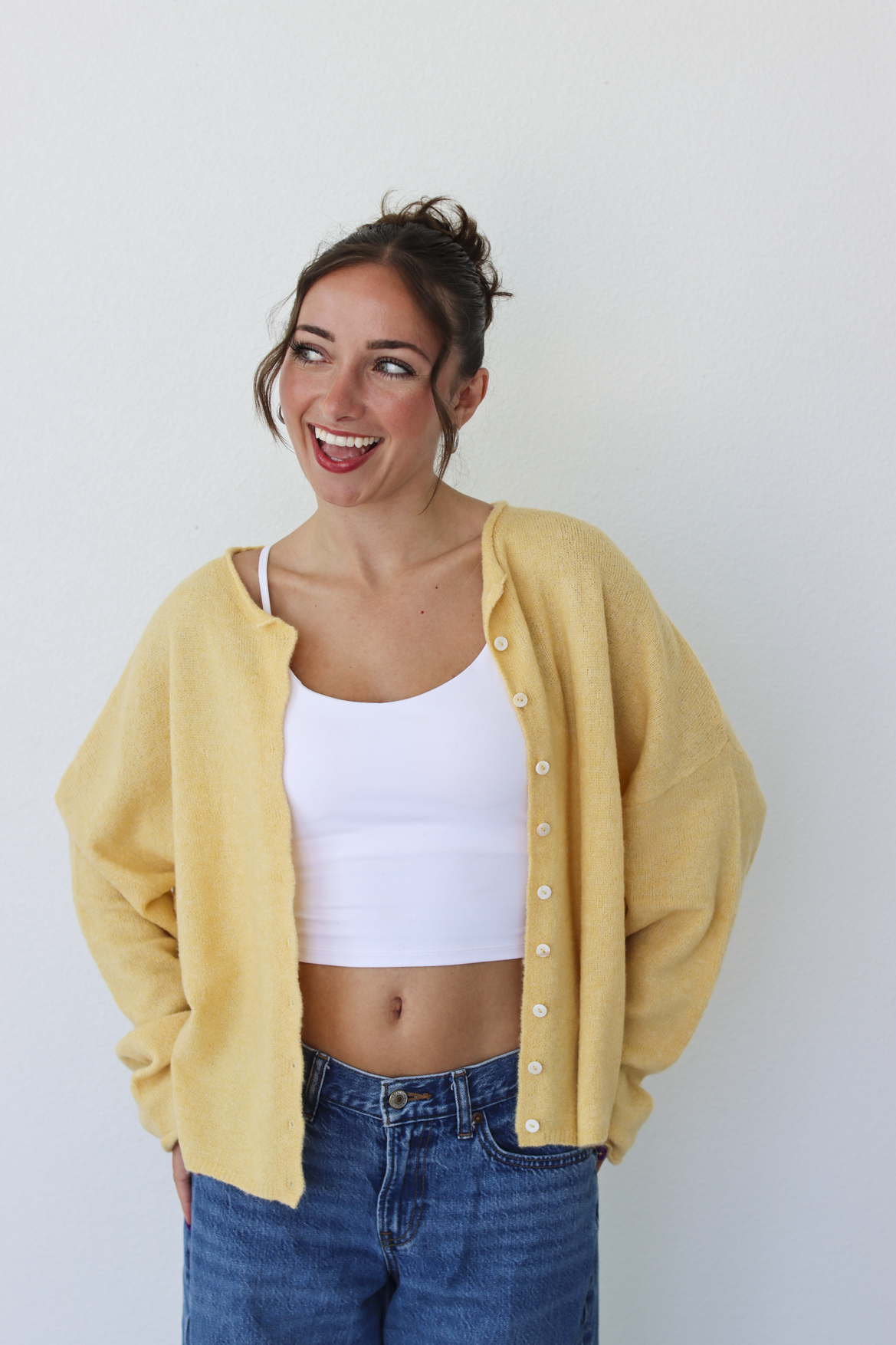 A woman smiling and wearing a yellow button-up cardigan over a white crop top and blue jeans, standing against a plain light gray wall.