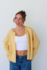 A woman smiling and wearing a yellow button-up cardigan over a white crop top and blue jeans, standing against a plain light gray wall.