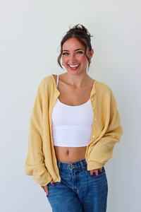 A woman smiling and wearing a yellow button-up cardigan over a white crop top and blue jeans, standing against a plain light gray wall.
