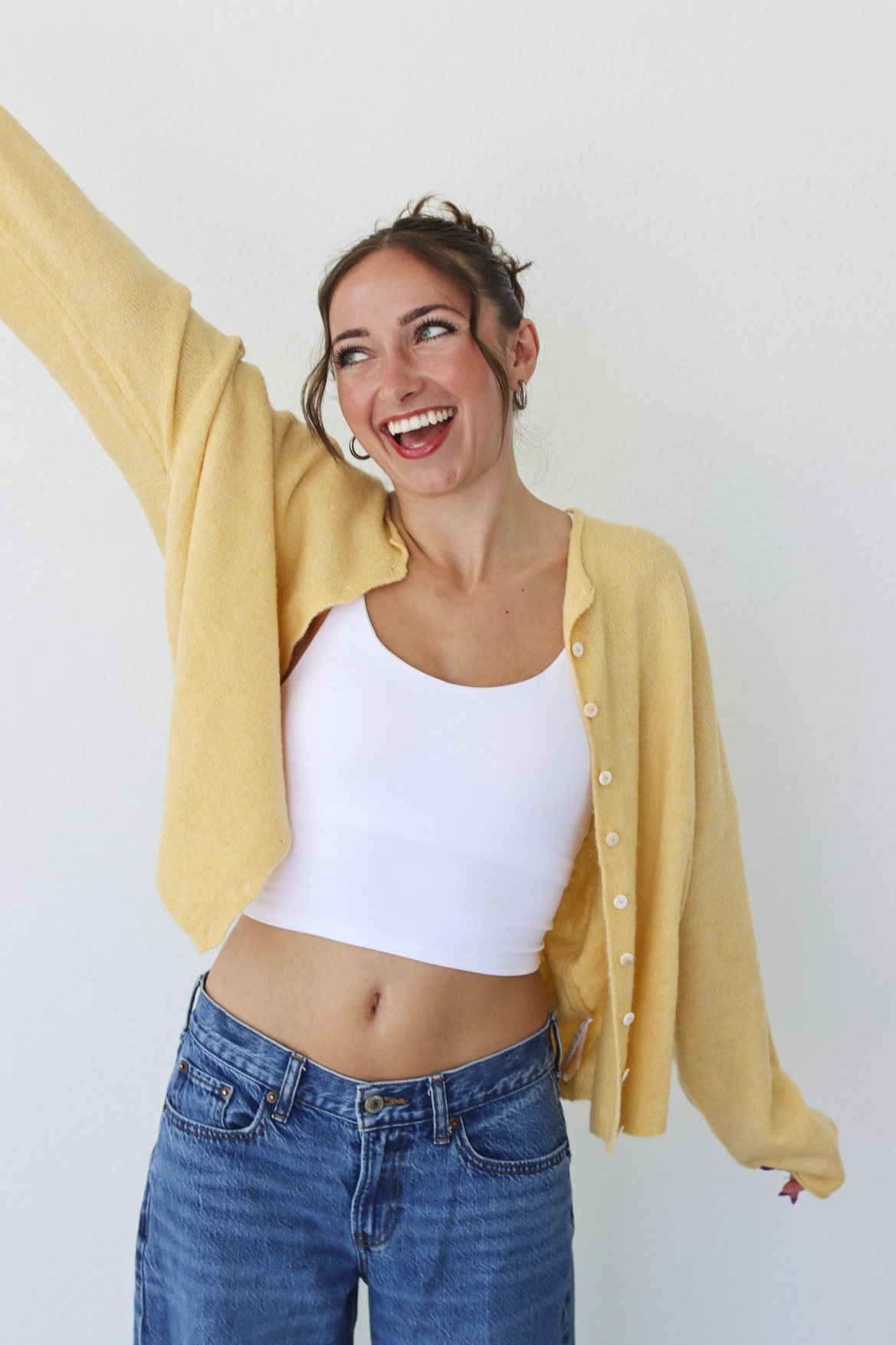 A woman smiling and wearing a yellow button-up cardigan over a white crop top and blue jeans, standing against a plain light gray wall.