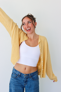 A woman smiling and wearing a yellow button-up cardigan over a white crop top and blue jeans, standing against a plain light gray wall.