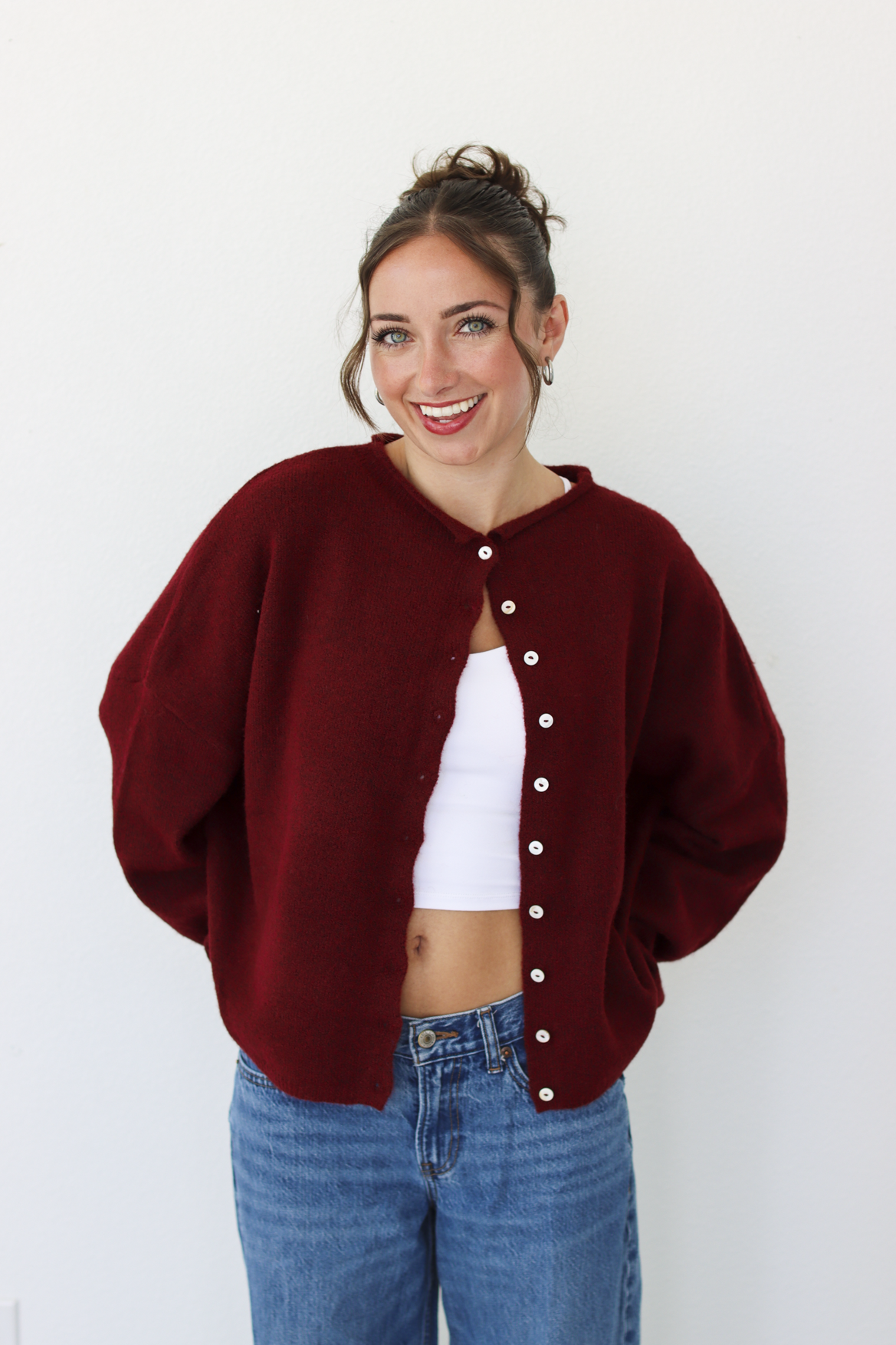 A woman wearing a burgundy knit cardigan with white buttons over a white crop top and blue jeans, standing against a plain white wall.