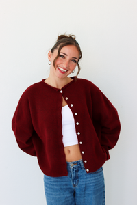 A woman wearing a burgundy knit cardigan with white buttons over a white crop top and blue jeans, standing against a plain white wall.