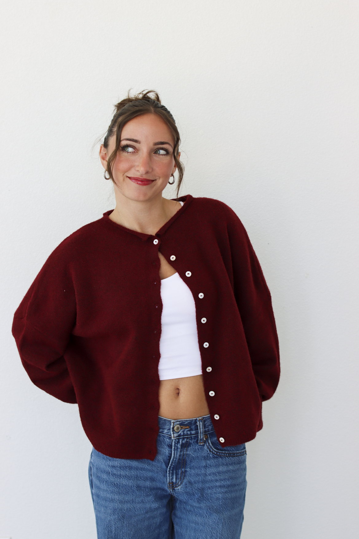 A woman wearing a burgundy knit cardigan with white buttons over a white crop top and blue jeans, standing against a plain white wall.