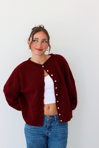 A woman wearing a burgundy knit cardigan with white buttons over a white crop top and blue jeans, standing against a plain white wall.