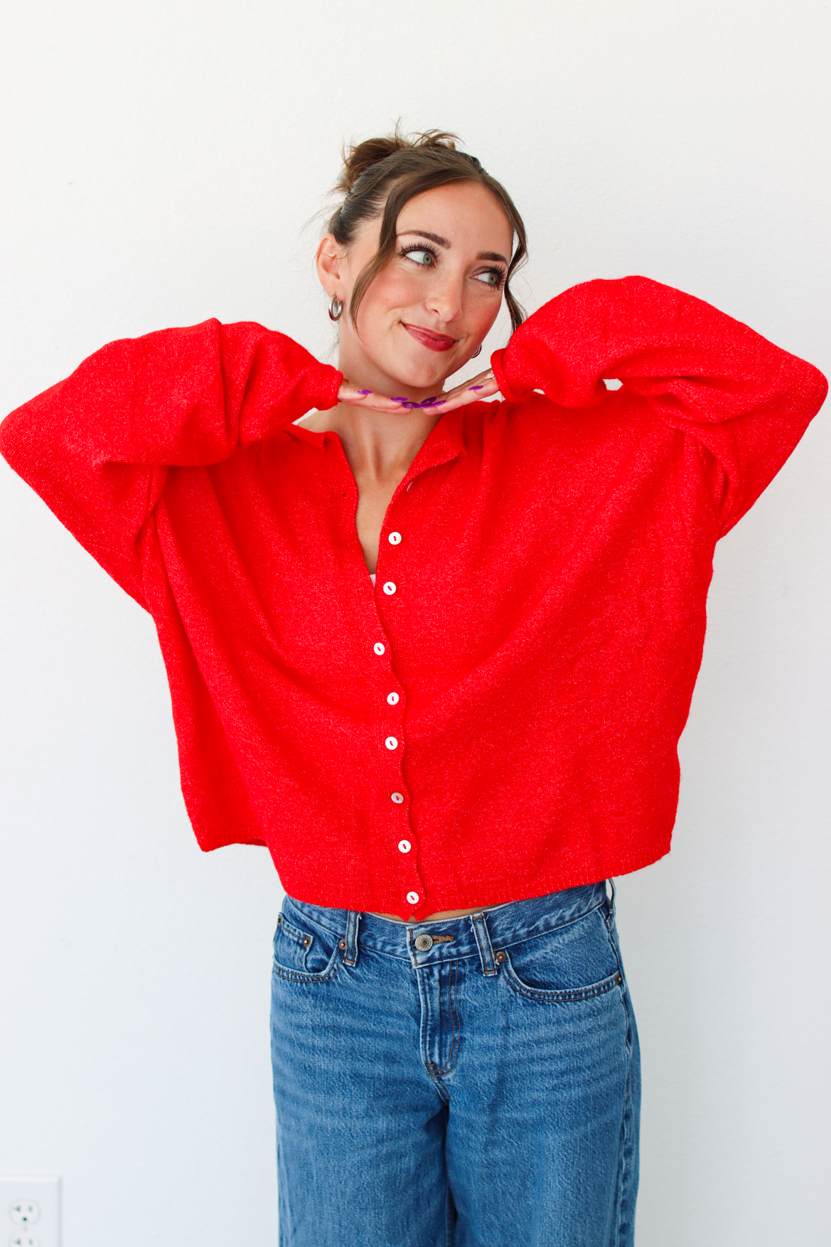A woman smiling and wearing a bright red knit button-front cardigan paired with blue jeans, standing against a plain white wall.