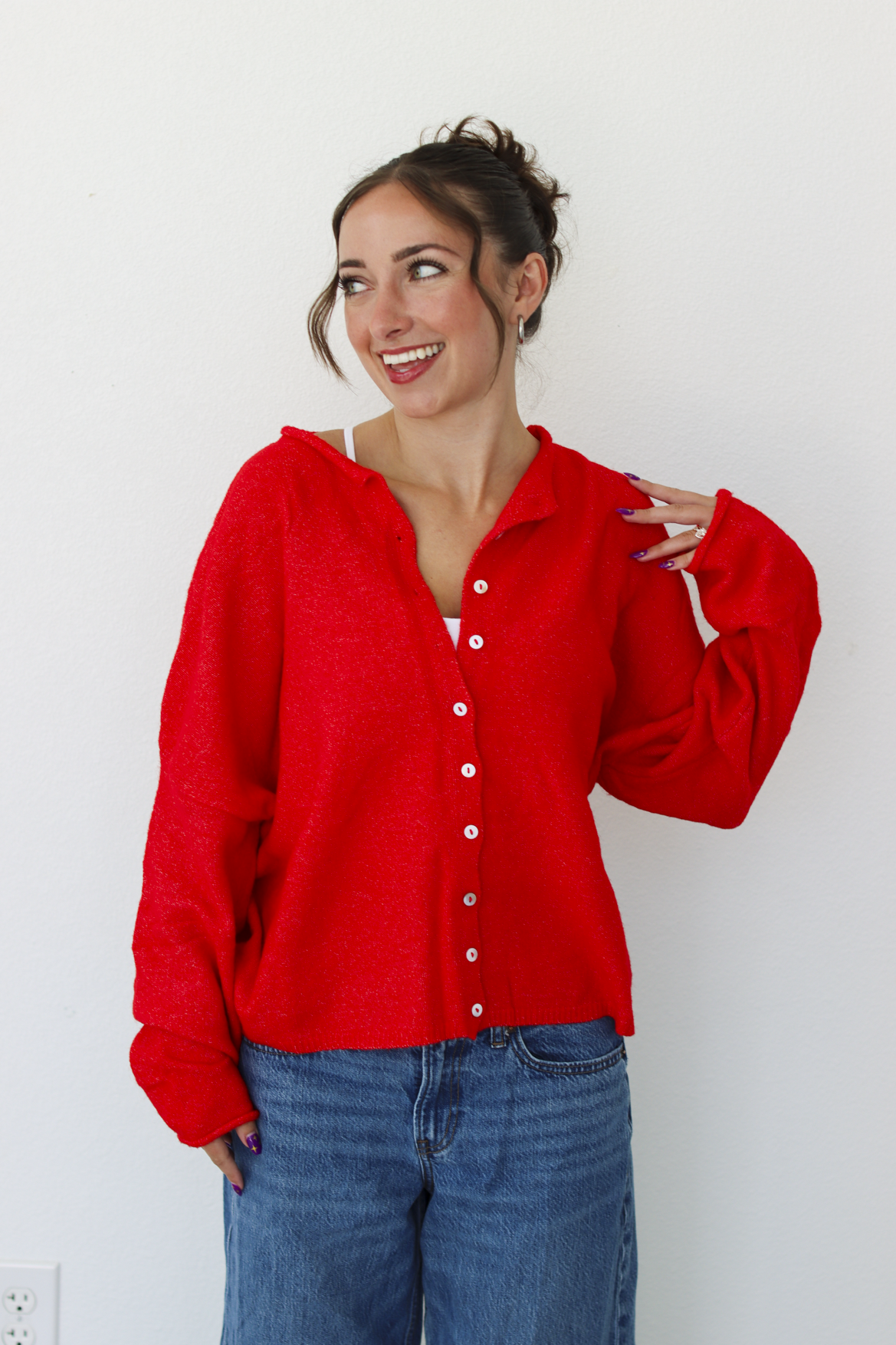 A woman smiling and wearing a bright red knit button-front cardigan paired with blue jeans, standing against a plain white wall.