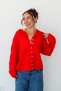 A woman smiling and wearing a bright red knit button-front cardigan paired with blue jeans, standing against a plain white wall.