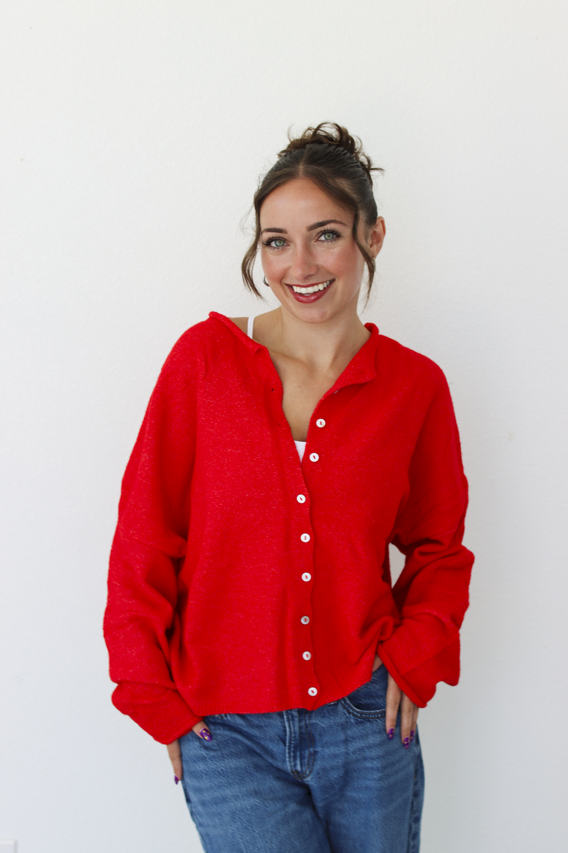 A woman smiling and wearing a bright red knit button-front cardigan paired with blue jeans, standing against a plain white wall.