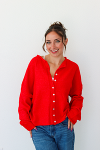 A woman smiling and wearing a bright red knit button-front cardigan paired with blue jeans, standing against a plain white wall.