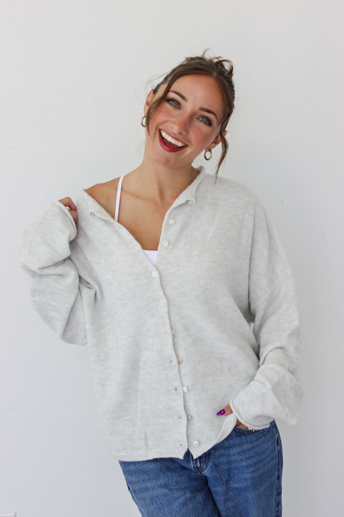 A woman smiling and wearing a light gray button-up cardigan over a white camisole and blue jeans, standing against a plain white wall.