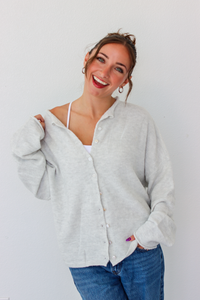 A woman smiling and wearing a light gray button-up cardigan over a white camisole and blue jeans, standing against a plain white wall.