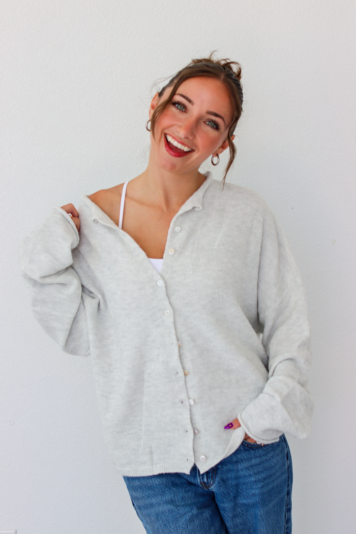 A woman smiling and wearing a light gray button-up cardigan over a white camisole and blue jeans, standing against a plain white wall.