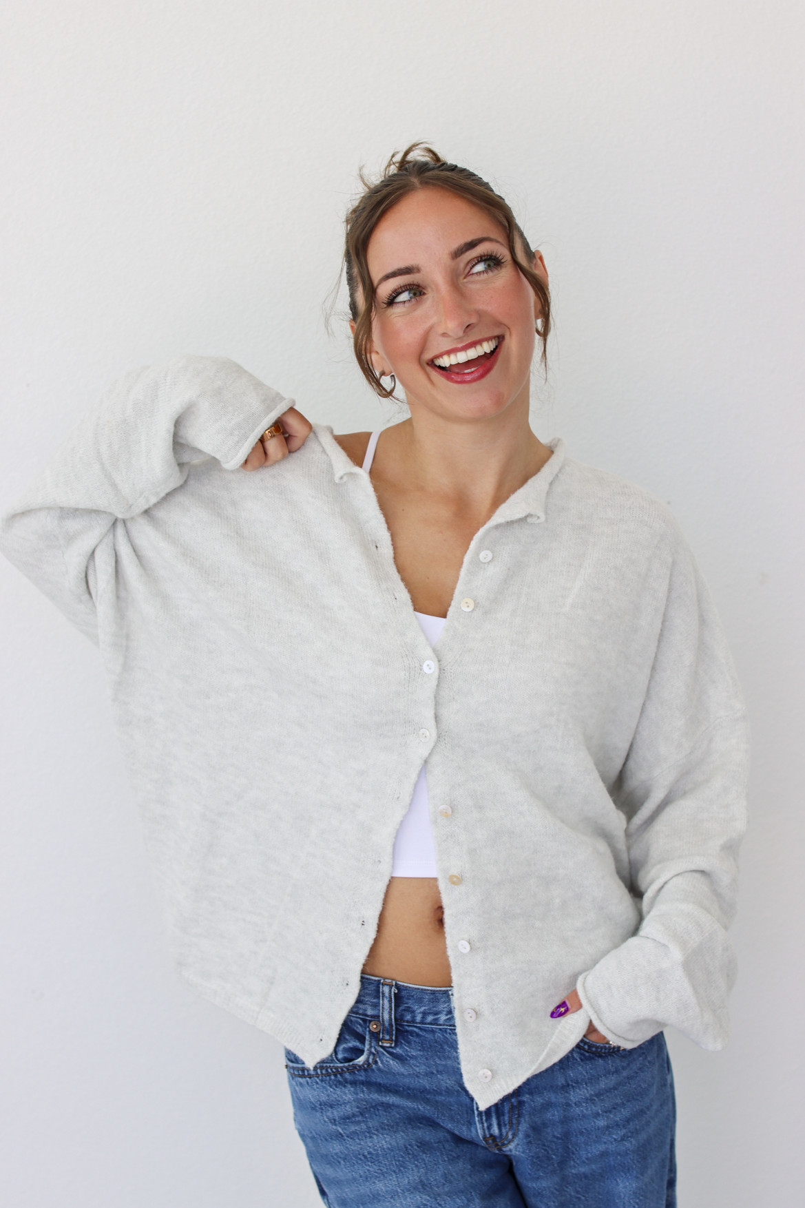 A woman smiling and wearing a light gray button-up cardigan over a white camisole and blue jeans, standing against a plain white wall.