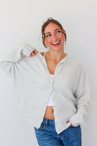 A woman smiling and wearing a light gray button-up cardigan over a white camisole and blue jeans, standing against a plain white wall.