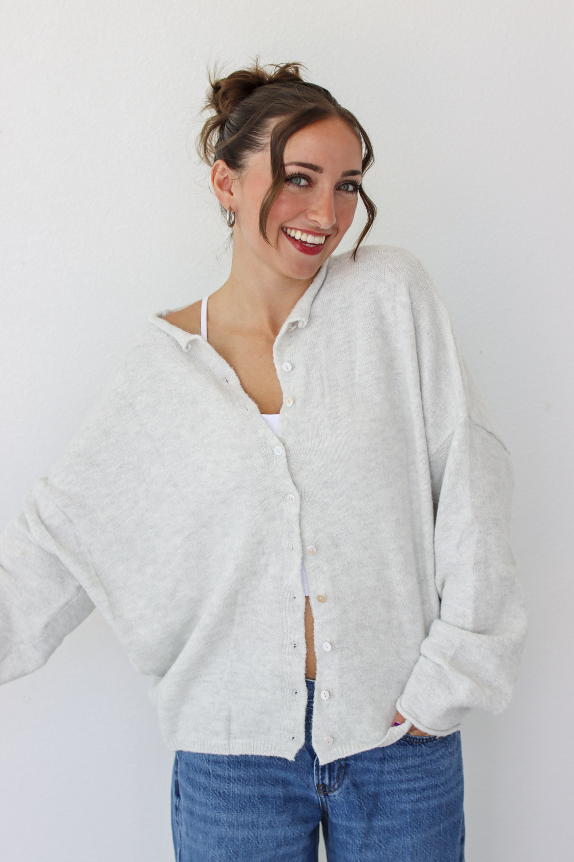 A woman smiling and wearing a light gray button-up cardigan over a white camisole and blue jeans, standing against a plain white wall.