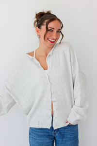 A woman smiling and wearing a light gray button-up cardigan over a white camisole and blue jeans, standing against a plain white wall.