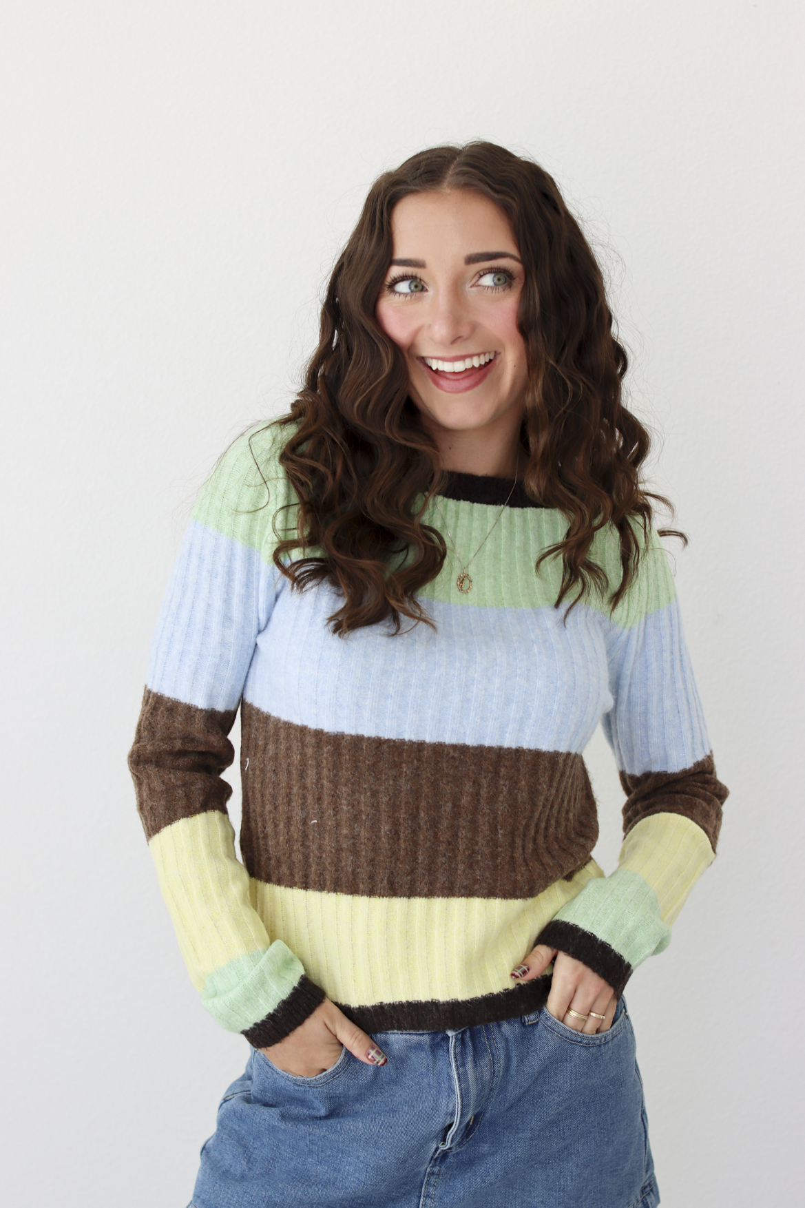 Woman wearing a long sleeve top with brown, green, blue, and yellow stripe and blue denim skirt. 