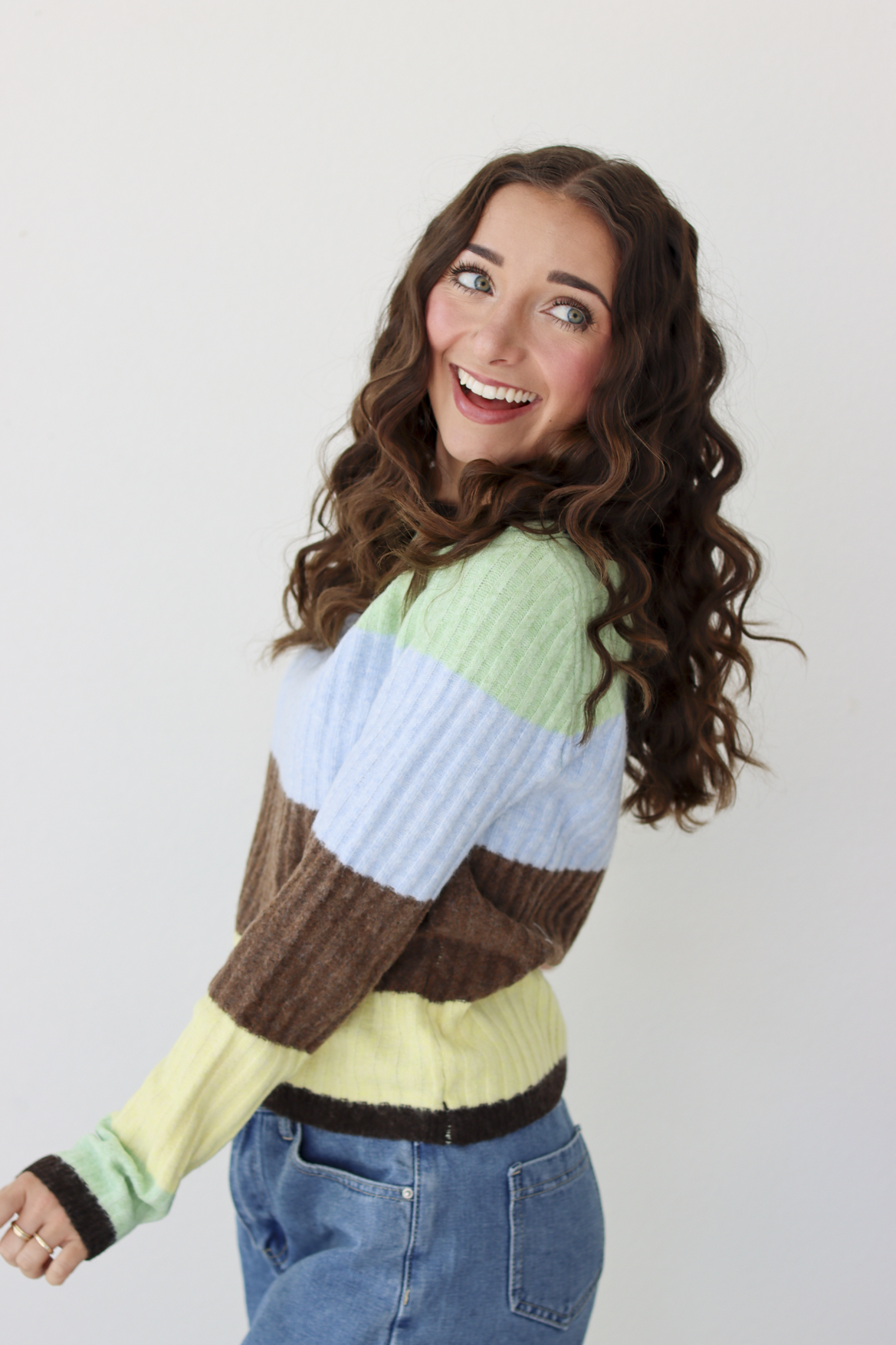 Woman wearing a long sleeve top with brown, green, blue, and yellow stripe and blue denim skirt. 