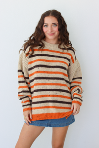 A woman wearing a chunky knit sweater with horizontal stripes in cream, orange, and dark brown, paired with a blue denim skirt, standing against a light gray background.