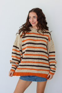 A woman wearing a chunky knit sweater with horizontal stripes in cream, orange, and dark brown, paired with a blue denim skirt, standing against a light gray background.
