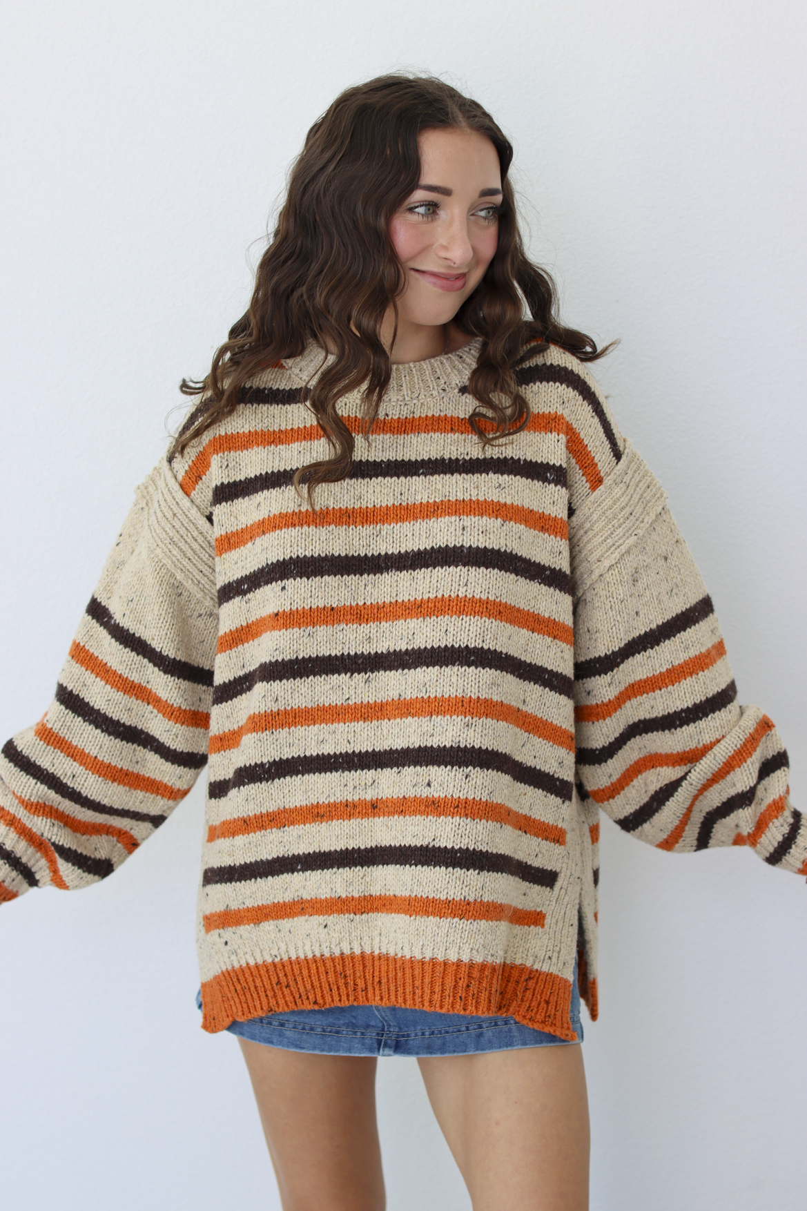 A woman wearing a chunky knit sweater with horizontal stripes in cream, orange, and dark brown, paired with a blue denim skirt, standing against a light gray background.