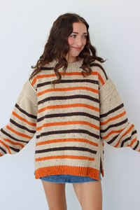 A woman wearing a chunky knit sweater with horizontal stripes in cream, orange, and dark brown, paired with a blue denim skirt, standing against a light gray background.