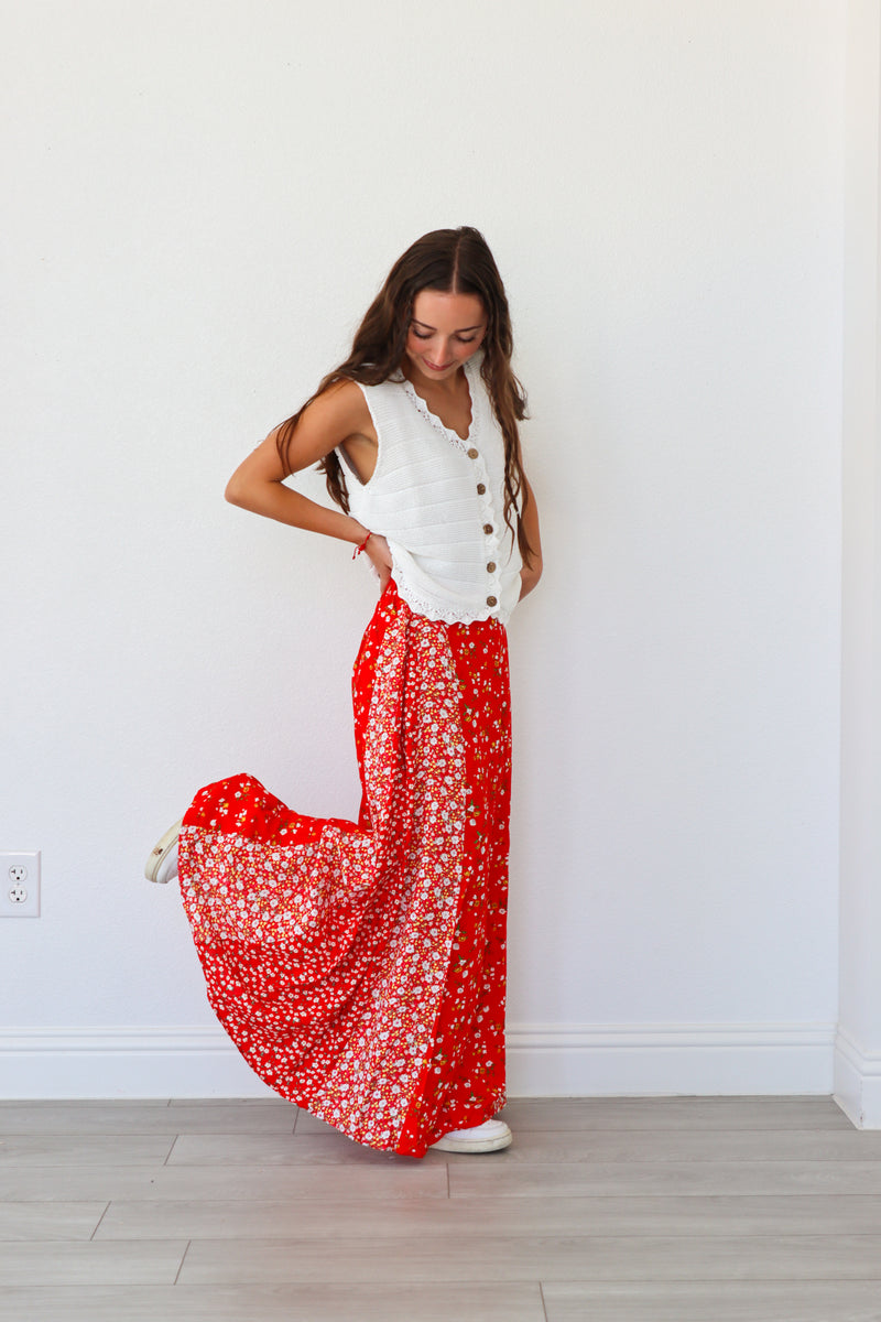 Woman wearing a white sleeveless top with buttons and a red floral skirt against a white wall.
