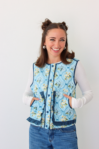 Woman wearing a blue floral puffer vest with a white long sleeve shirt and blue denim jeans 
