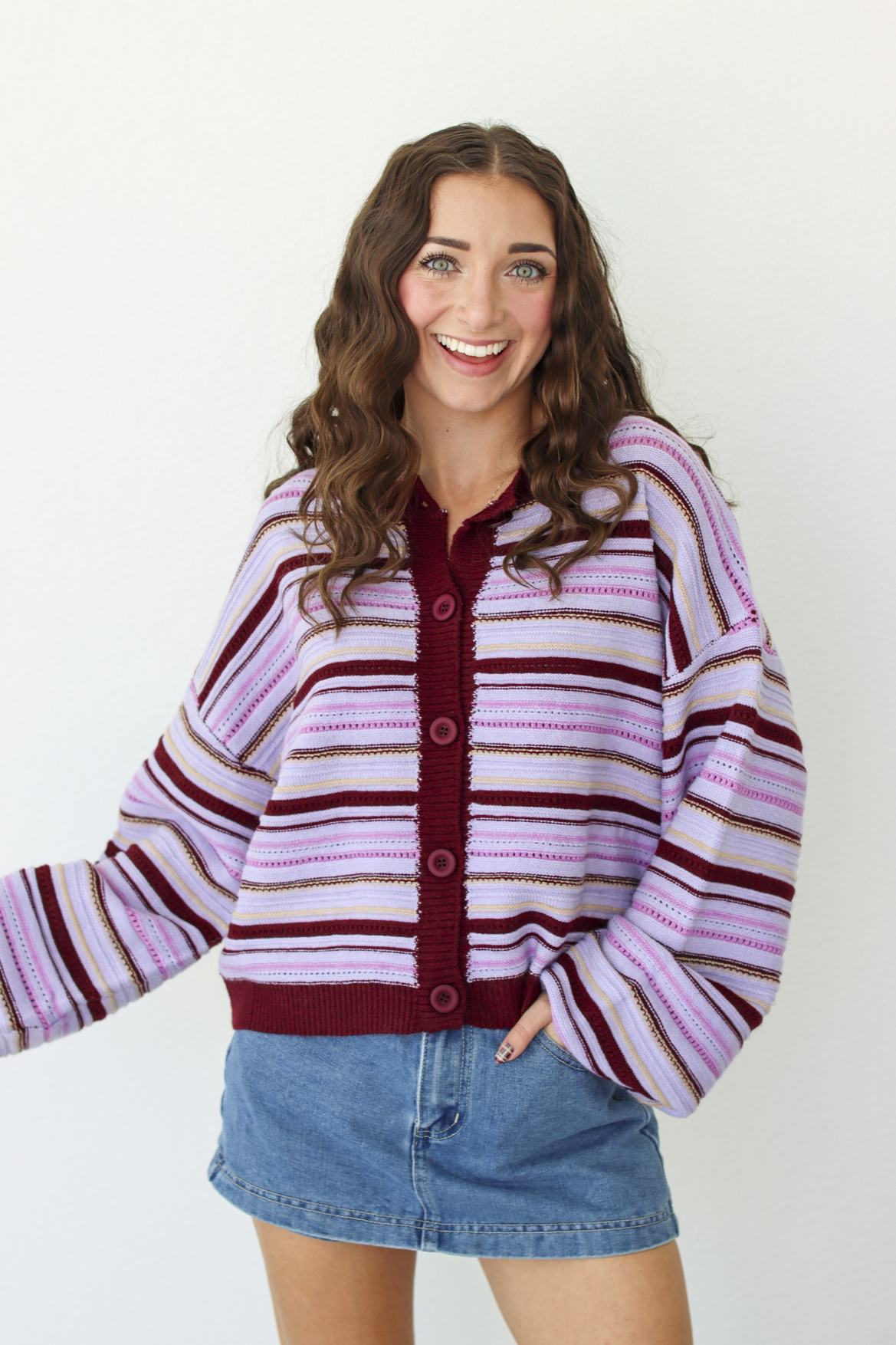 A woman modeling a pink and dark purple striped knit cardigan with a button-front closure, paired with a denim mini skirt, against a plain white background.