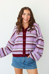 A woman modeling a pink and dark purple striped knit cardigan with a button-front closure, paired with a denim mini skirt, against a plain white background.