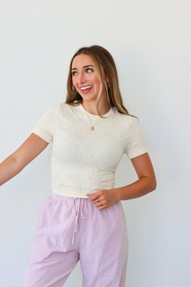 Woman wearing a white textured top and light purple pants against a white background