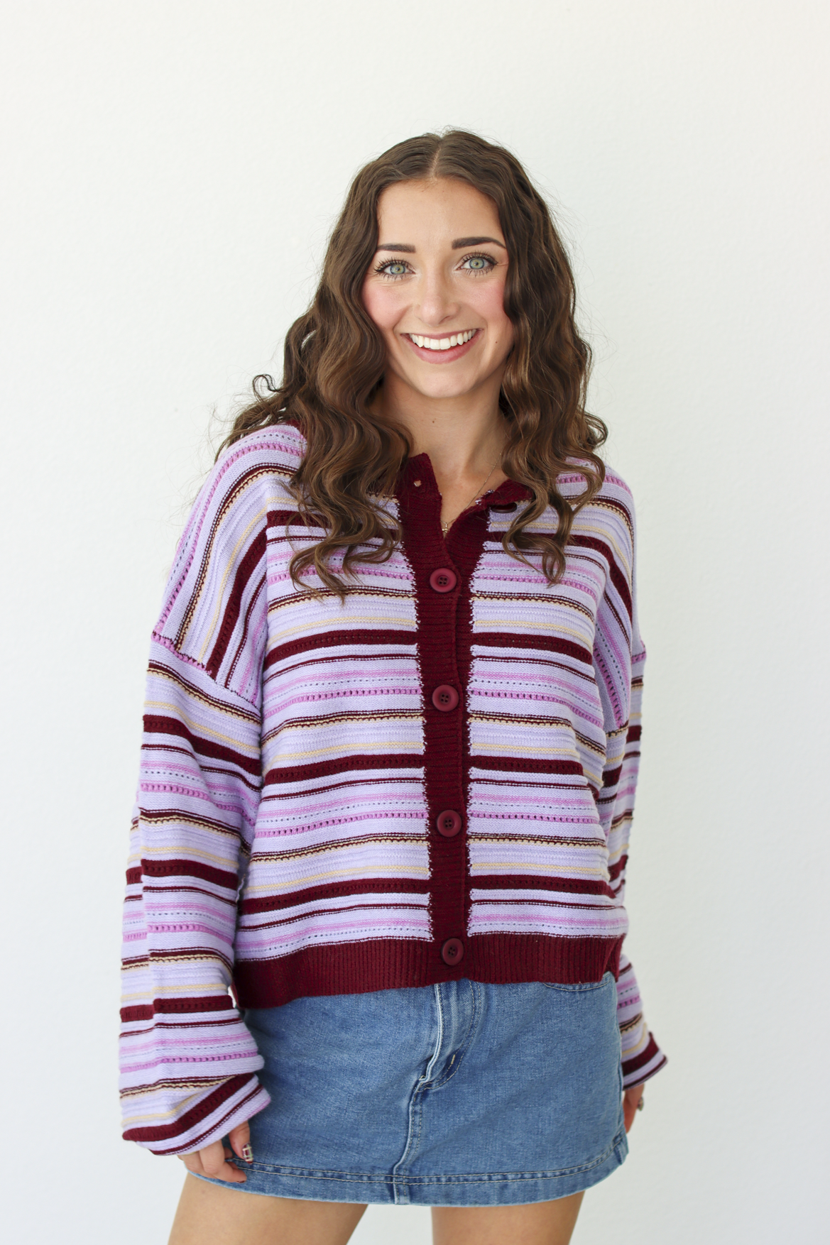A woman with long brown hair smiling, wearing a lavender and dark purple striped knit cardigan with a button-up front, paired with a blue denim mini skirt, against a plain white background.