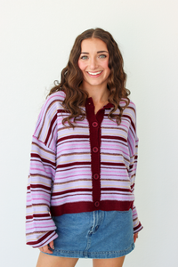 A woman with long brown hair smiling, wearing a lavender and dark purple striped knit cardigan with a button-up front, paired with a blue denim mini skirt, against a plain white background.